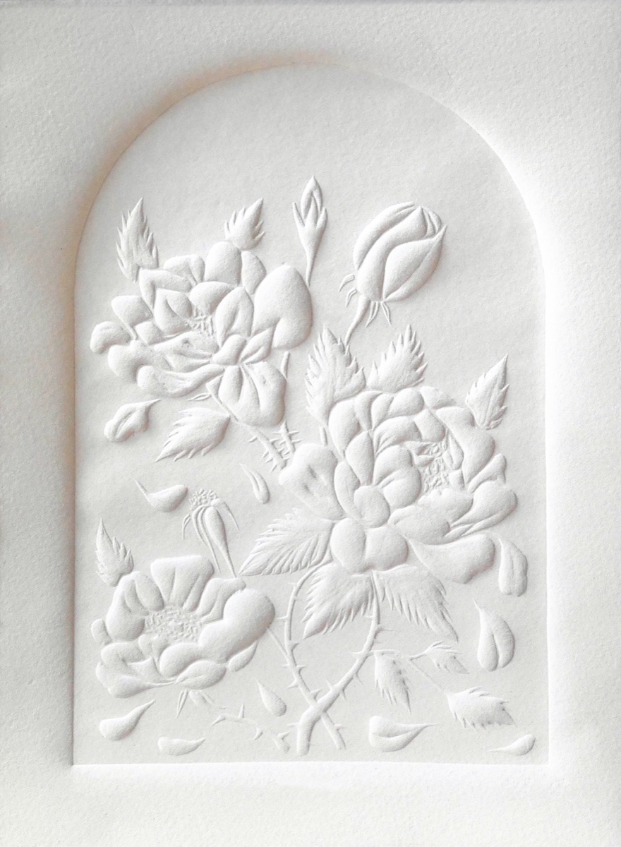 Roses, buds, thorned stems and leaves inside an arched window, every petal standing up from white paper with no ink, and loose petals falling around them. She carved it beside a bouquet cut from her own garden, coming back each morning to find more petals on the desk, and chose to show the flowers as they let go rather than in full bloom.